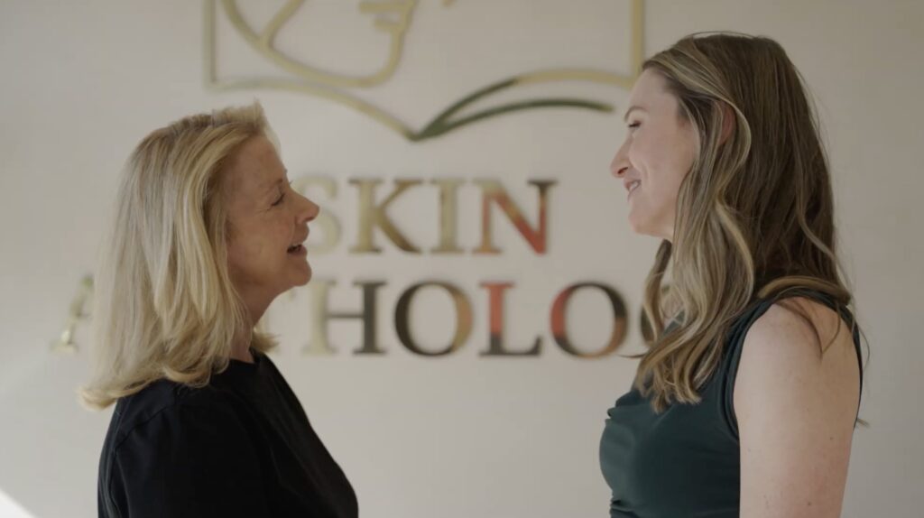 A client and a Skin Anthology provider talking in front of the gold Skin Anthology logo wall